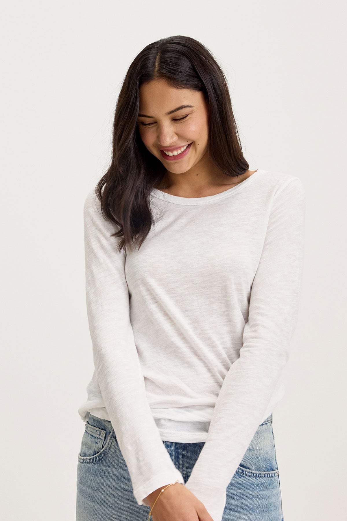   A person with long dark hair smiles while looking down, wearing the LIZZIE TEE by Velvet by Graham & Spencer and blue jeans, standing against a plain white background. 