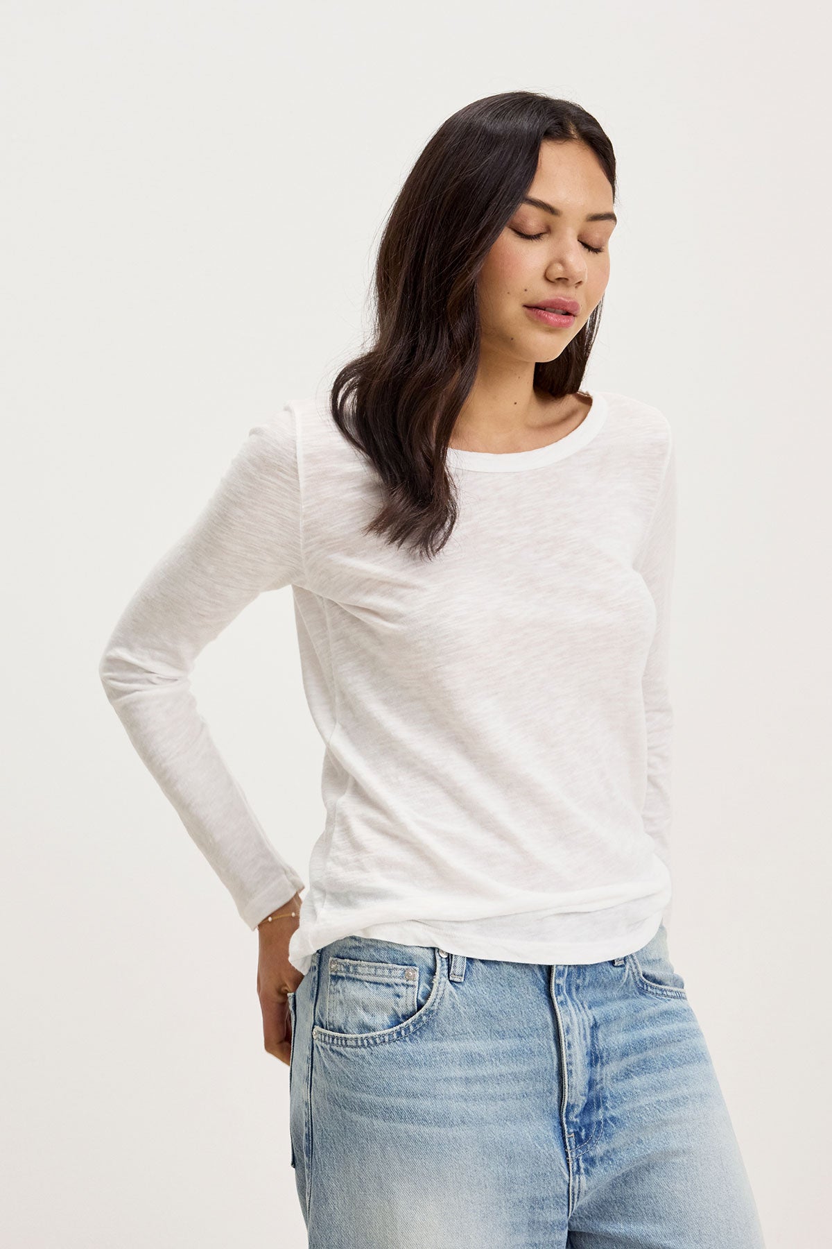   A woman wearing the LIZZIE TEE by Velvet by Graham & Spencer, a white long-sleeve shirt with a classic crew neckline, and blue jeans stands with her eyes closed, hands resting on her hips. 