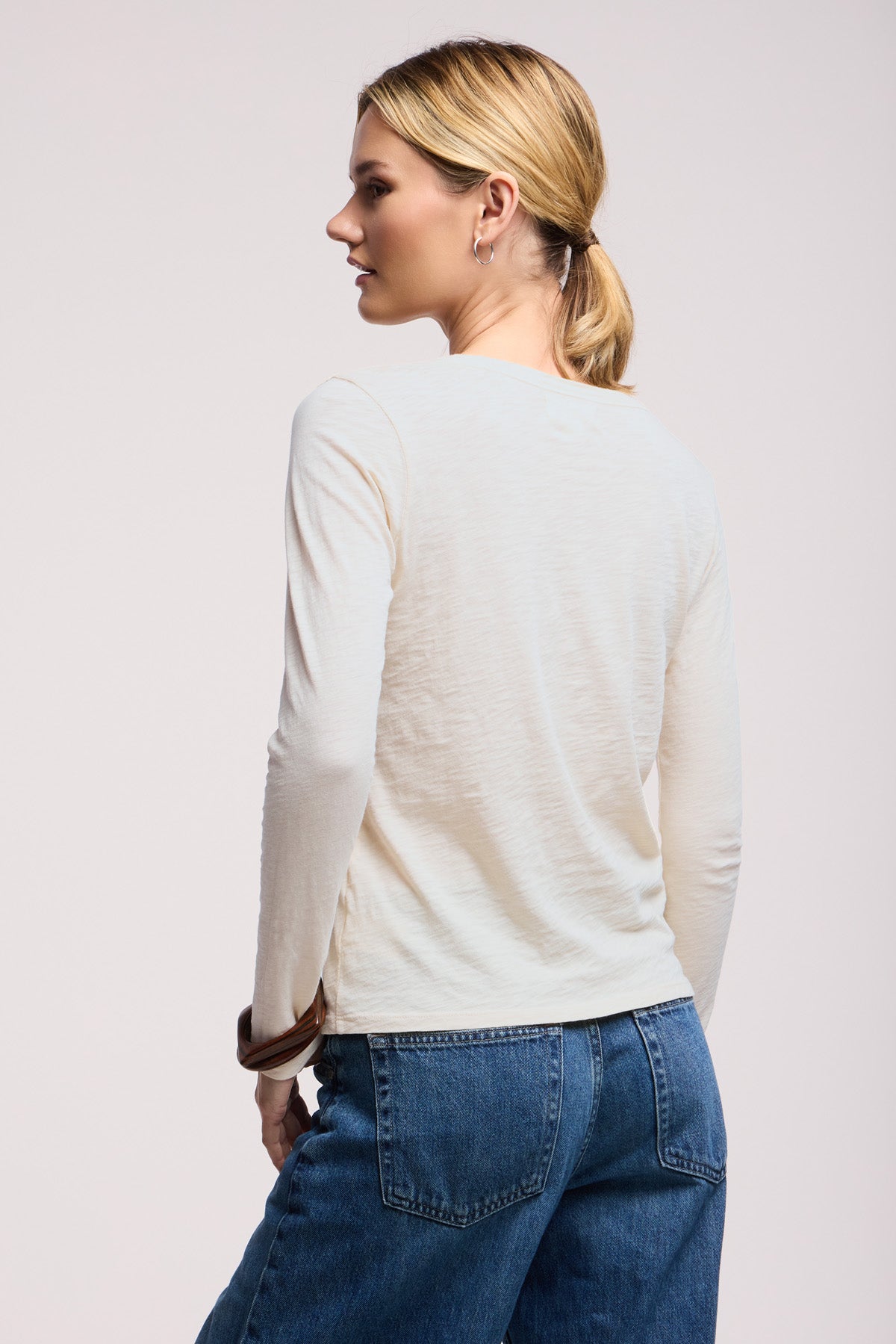 A woman with blonde hair in a low ponytail is seen from the back, wearing the LIZZIE TEE by Velvet by Graham & Spencer and blue jeans, standing against a plain light background.-41629819961537
