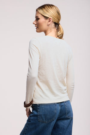 A woman with blonde hair in a low ponytail is seen from the back, wearing the LIZZIE TEE by Velvet by Graham & Spencer and blue jeans, standing against a plain light background.