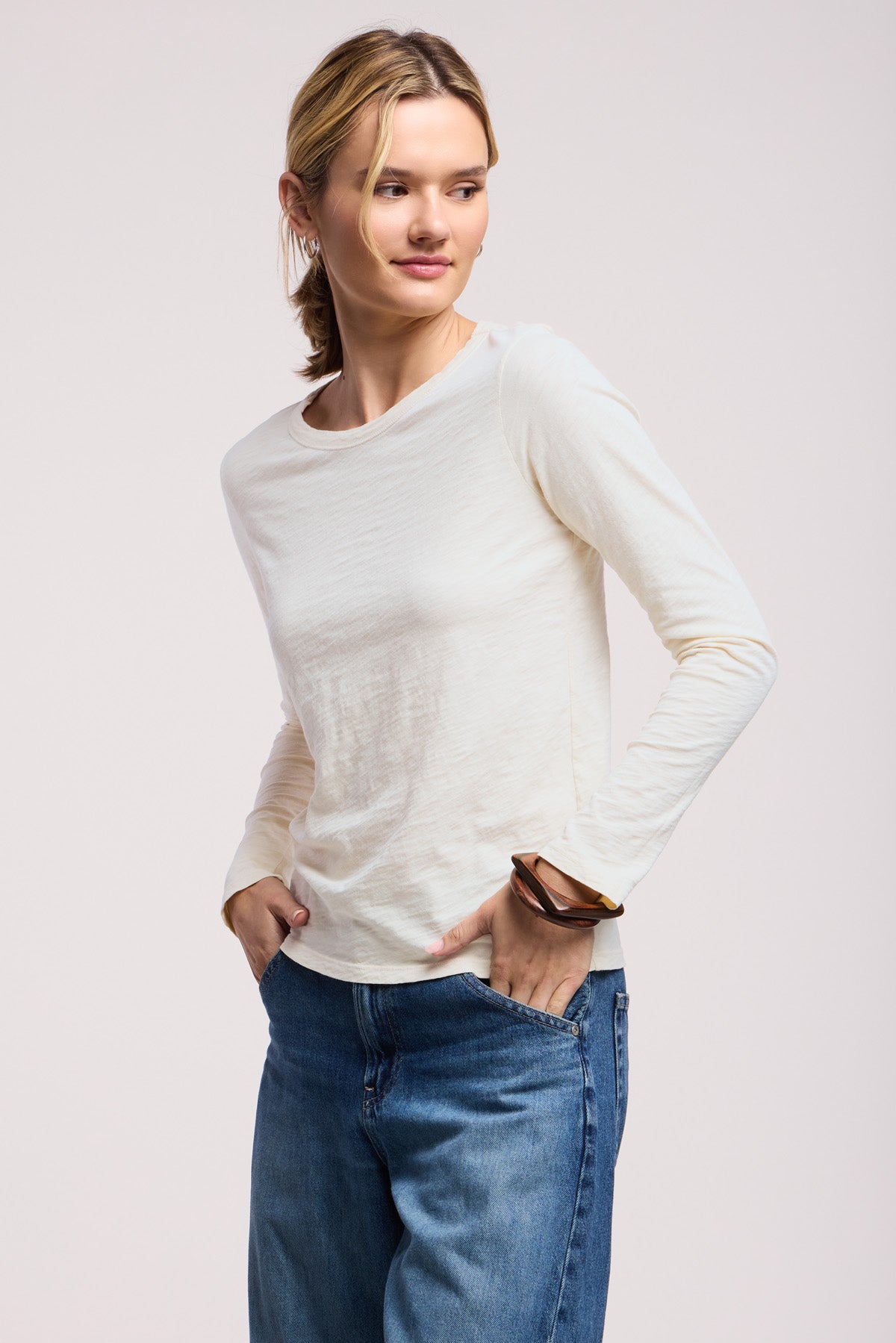 A person wears the LIZZIE TEE by Velvet by Graham & Spencer, a soft cream crew neck long sleeve top, paired with blue jeans, standing with hands in pockets against a plain background.-41629819994305