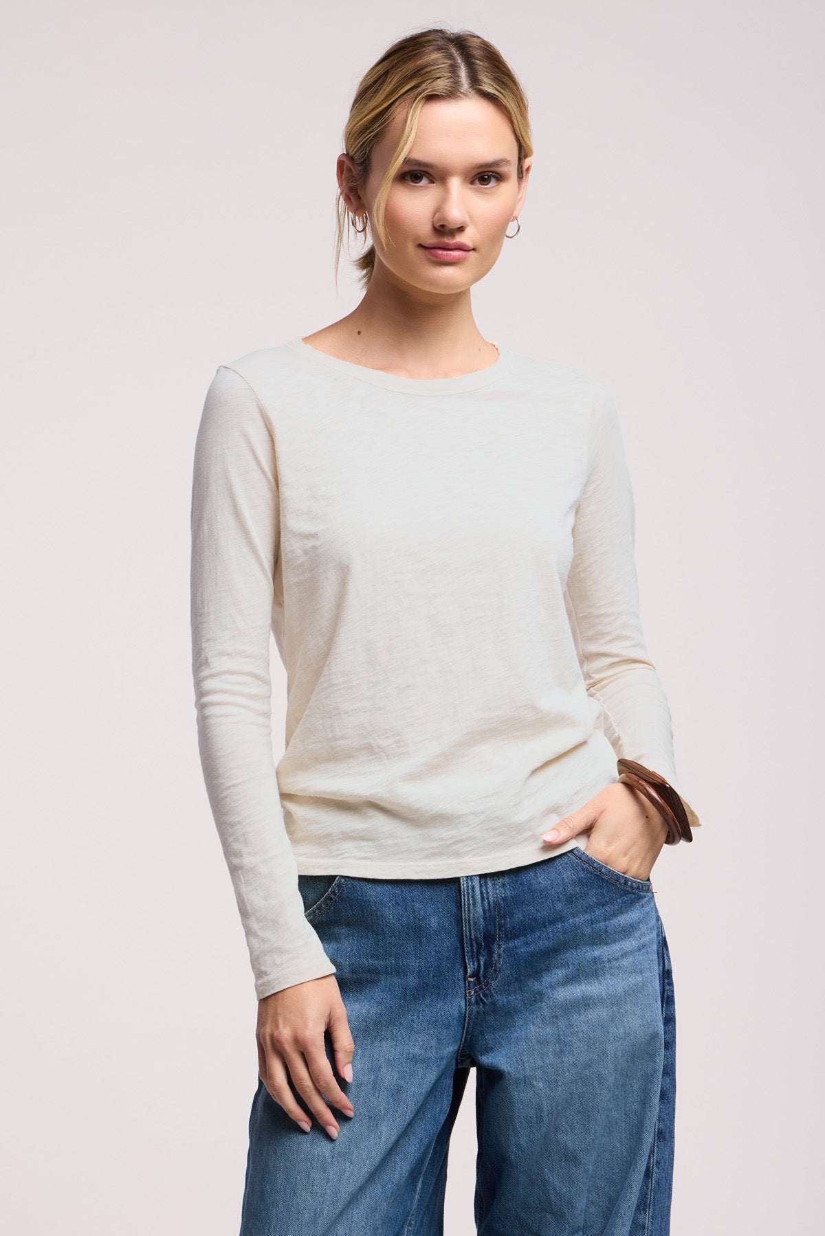 Someone stands facing forward against a plain background, wearing the LIZZIE TEE—a long-sleeve cotton slub crewneck from Velvet by Graham & Spencer—paired with blue jeans, one hand in their pocket.-41629820027073