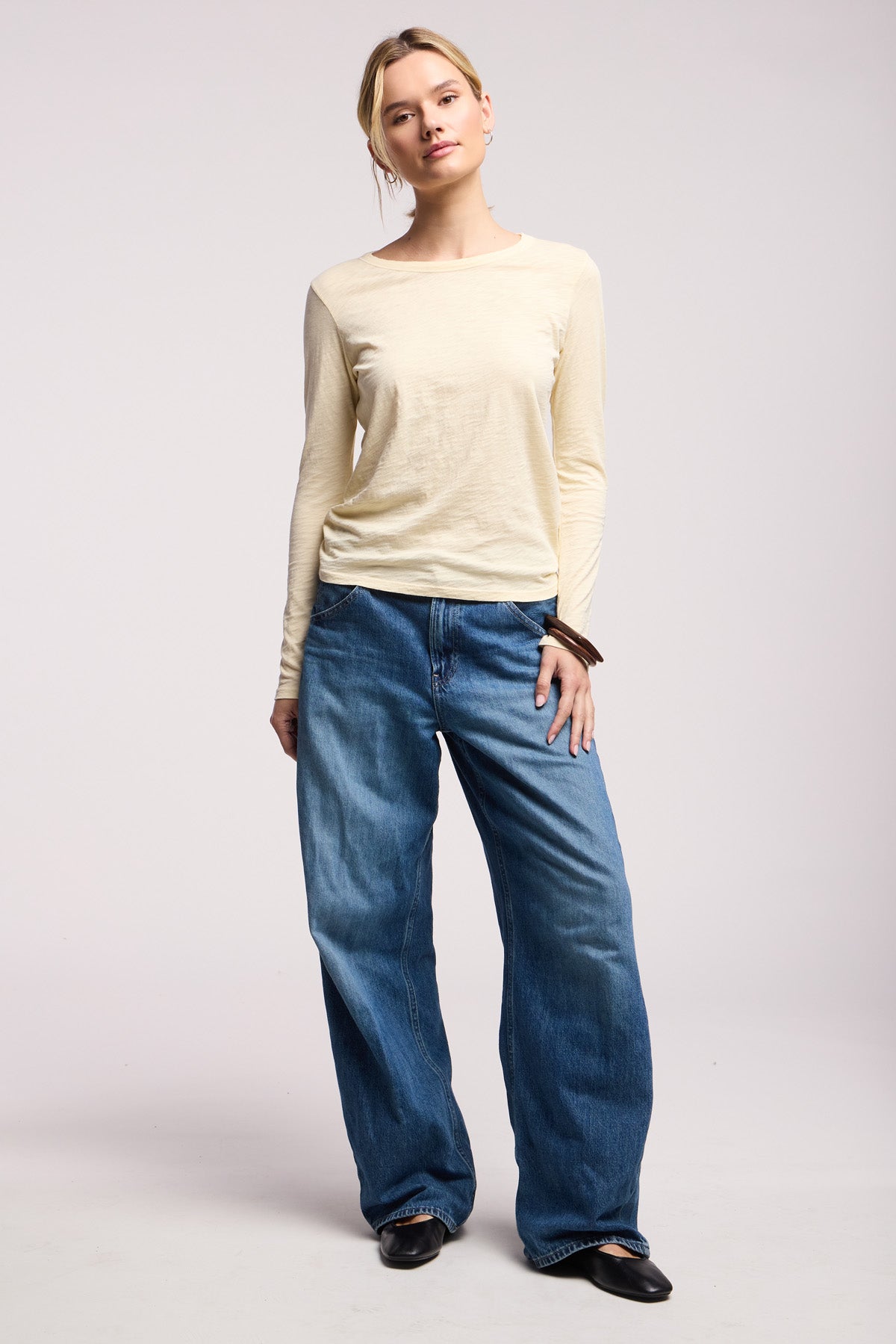 Someone stands against a plain background, wearing the LIZZIE TEE by Velvet by Graham & Spencer in beige, paired with loose-fitting blue jeans and black shoes.-40521694642369