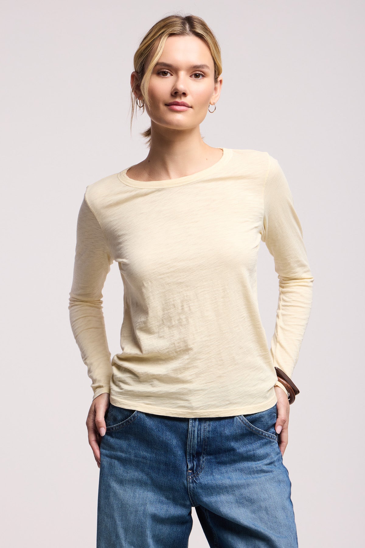 A person wearing the LIZZIE TEE by Velvet by Graham & Spencer, a light yellow cotton slub long sleeve crew neck top, pairs it with blue jeans and stands with hands in pockets against a plain background.