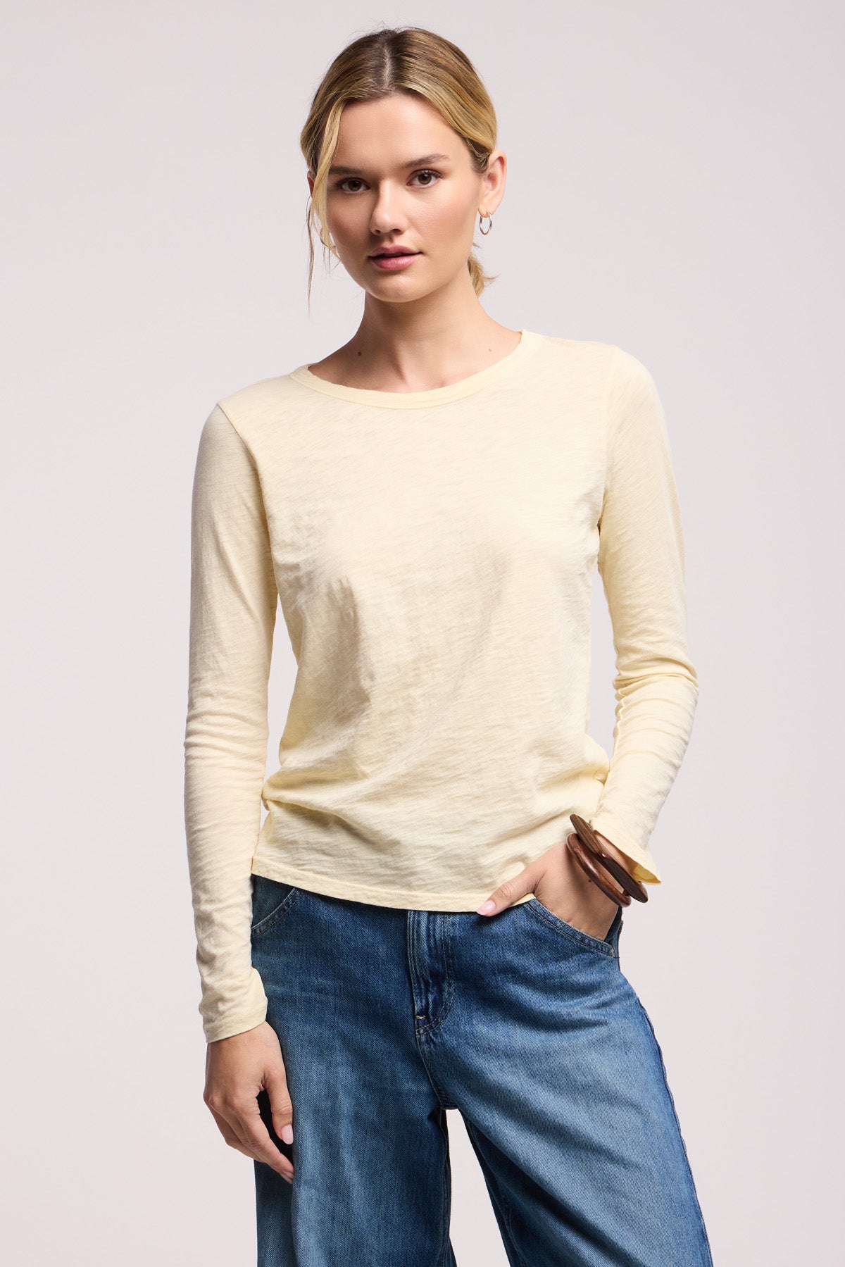 A woman faces forward against a plain background, wearing the Velvet by Graham & Spencer LIZZIE TEE—a light yellow cotton slub long sleeve crewneck—and blue jeans, with one hand in her pocket.