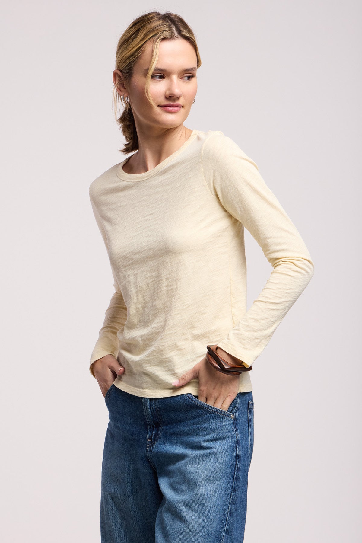 The woman wears the LIZZIE TEE by Velvet by Graham & Spencer, a light beige textured cotton slub long sleeve crewneck, paired with blue jeans. She stands with hands in pockets, looking to the side against a plain background.-40521694380225