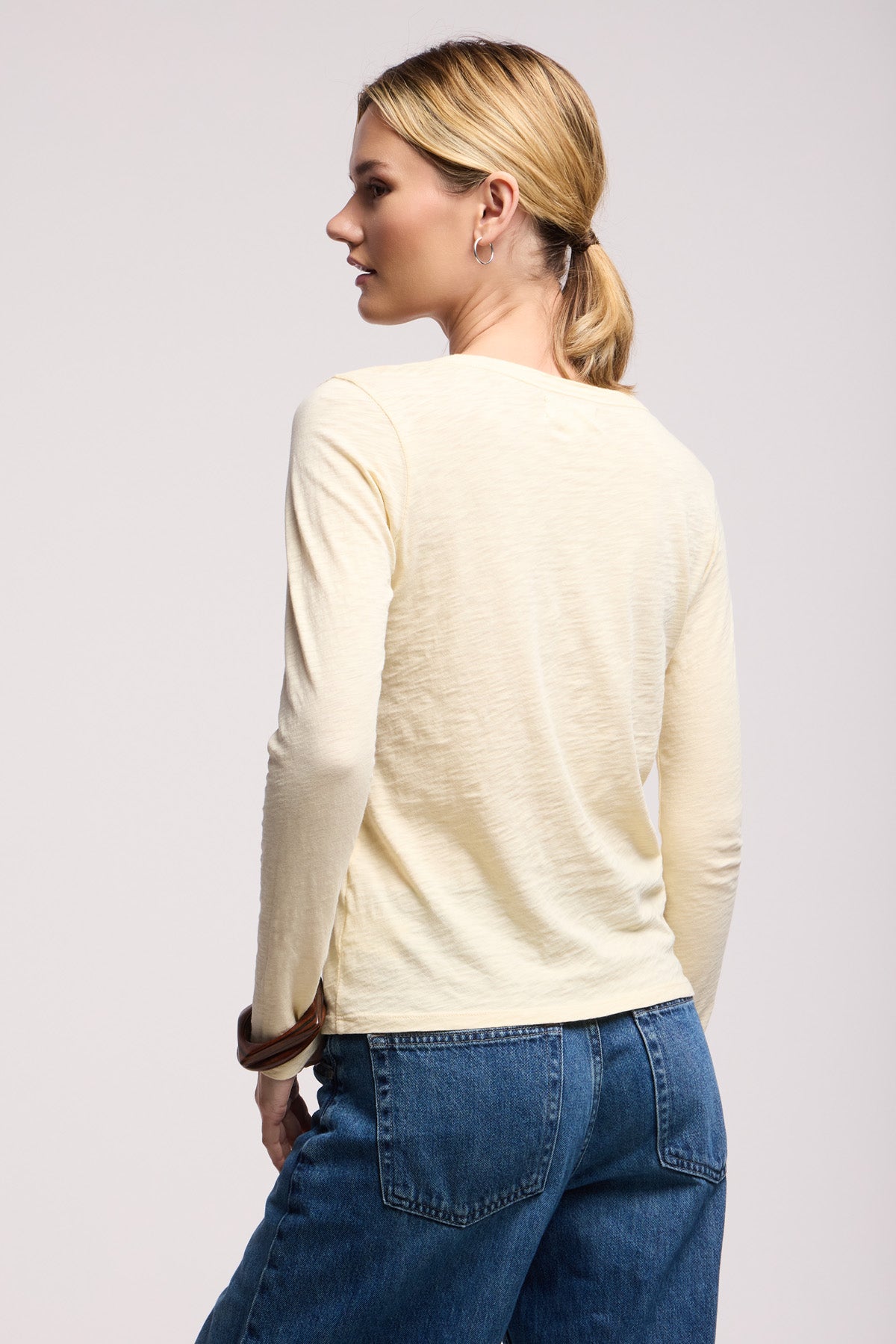 A woman with blond hair in a low ponytail is shown from the back, wearing the LIZZIE TEE by Velvet by Graham & Spencer—a cream long sleeve crewneck—paired with blue jeans against a plain background.