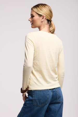 A woman with blond hair in a low ponytail is shown from the back, wearing the LIZZIE TEE by Velvet by Graham & Spencer—a cream long sleeve crewneck—paired with blue jeans against a plain background.