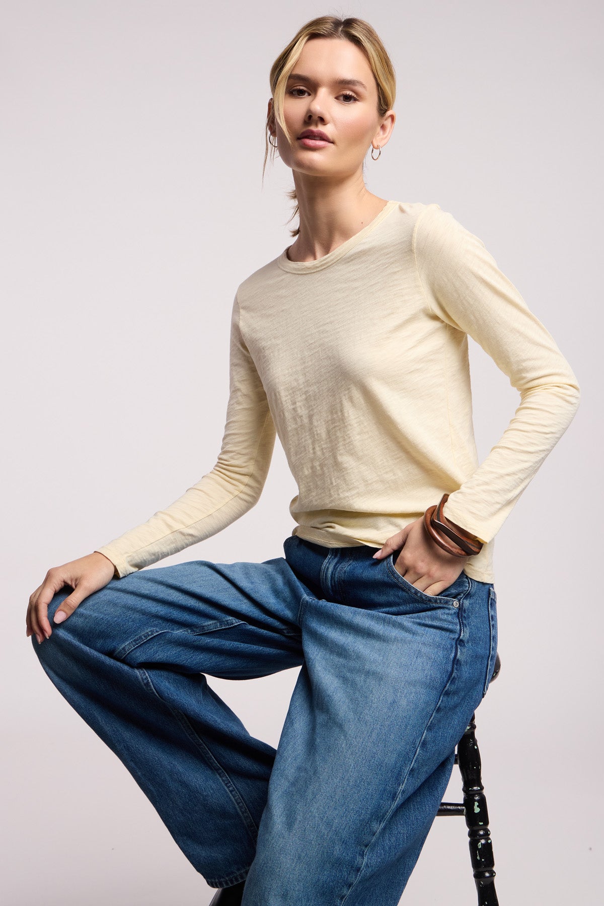 A person wears the LIZZIE TEE by Velvet by Graham & Spencer—a light yellow cotton slub long sleeve crewneck—paired with wide-leg blue jeans, sitting on a stool against a plain background.-40521694281921