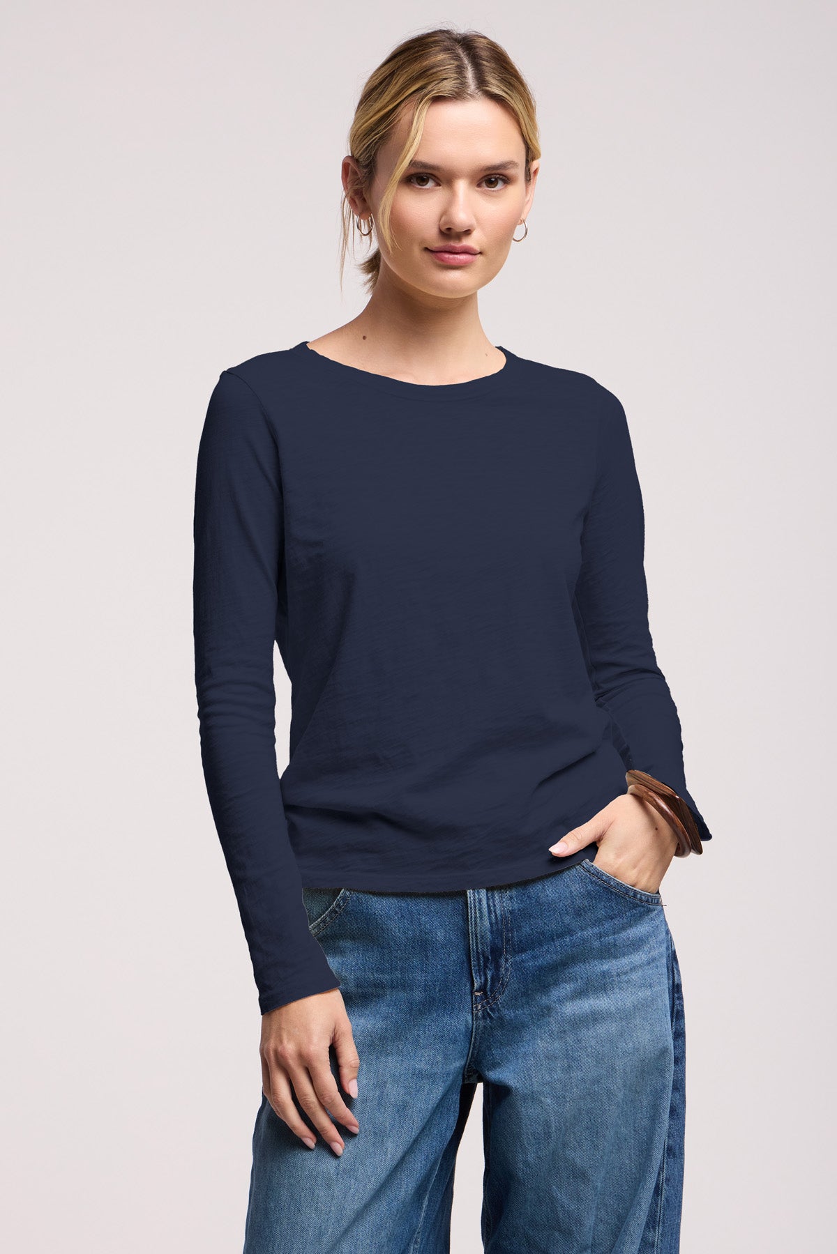 A woman wears the LIZZIE TEE by Velvet by Graham & Spencer, a long sleeve cotton slub top with a crew neckline, paired with blue jeans, standing with one hand in her pocket against a plain background.-41629820059841