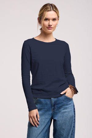 A woman wears the LIZZIE TEE by Velvet by Graham & Spencer, a long sleeve cotton slub top with a crew neckline, paired with blue jeans, standing with one hand in her pocket against a plain background.