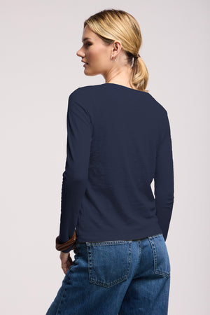 A woman with blonde hair in a low ponytail is seen from the back against a plain light background, wearing the LIZZIE TEE by Velvet by Graham & Spencer and blue jeans.