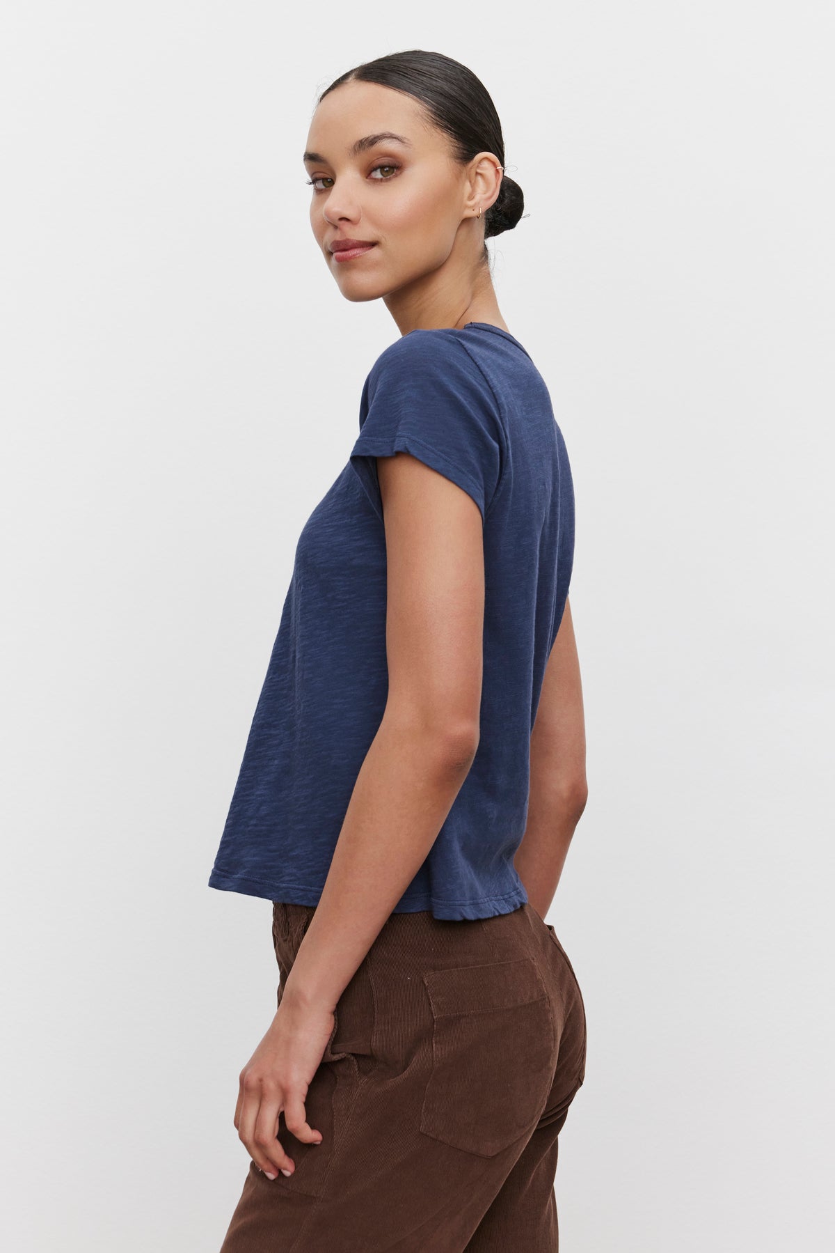   A person wearing a blue MARIE TEE by Velvet by Graham & Spencer with a boxy fit and brown pants stands against a white background, facing slightly to the side, perfect for a casual occasion. 