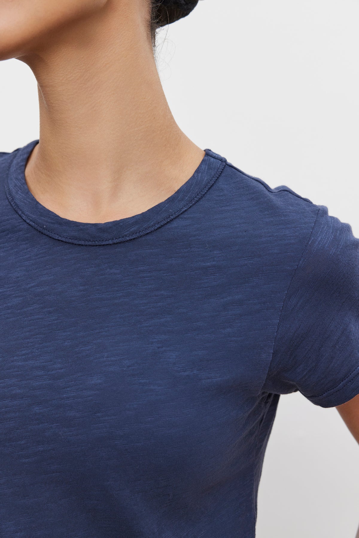   Close-up of a person wearing the MARIE TEE by Velvet by Graham & Spencer, a textured navy blue cotton slub T-shirt, with their head turned away from the camera. The boxy fit adds to the relaxed vibe, perfect for any casual occasion. The background is plain white. 