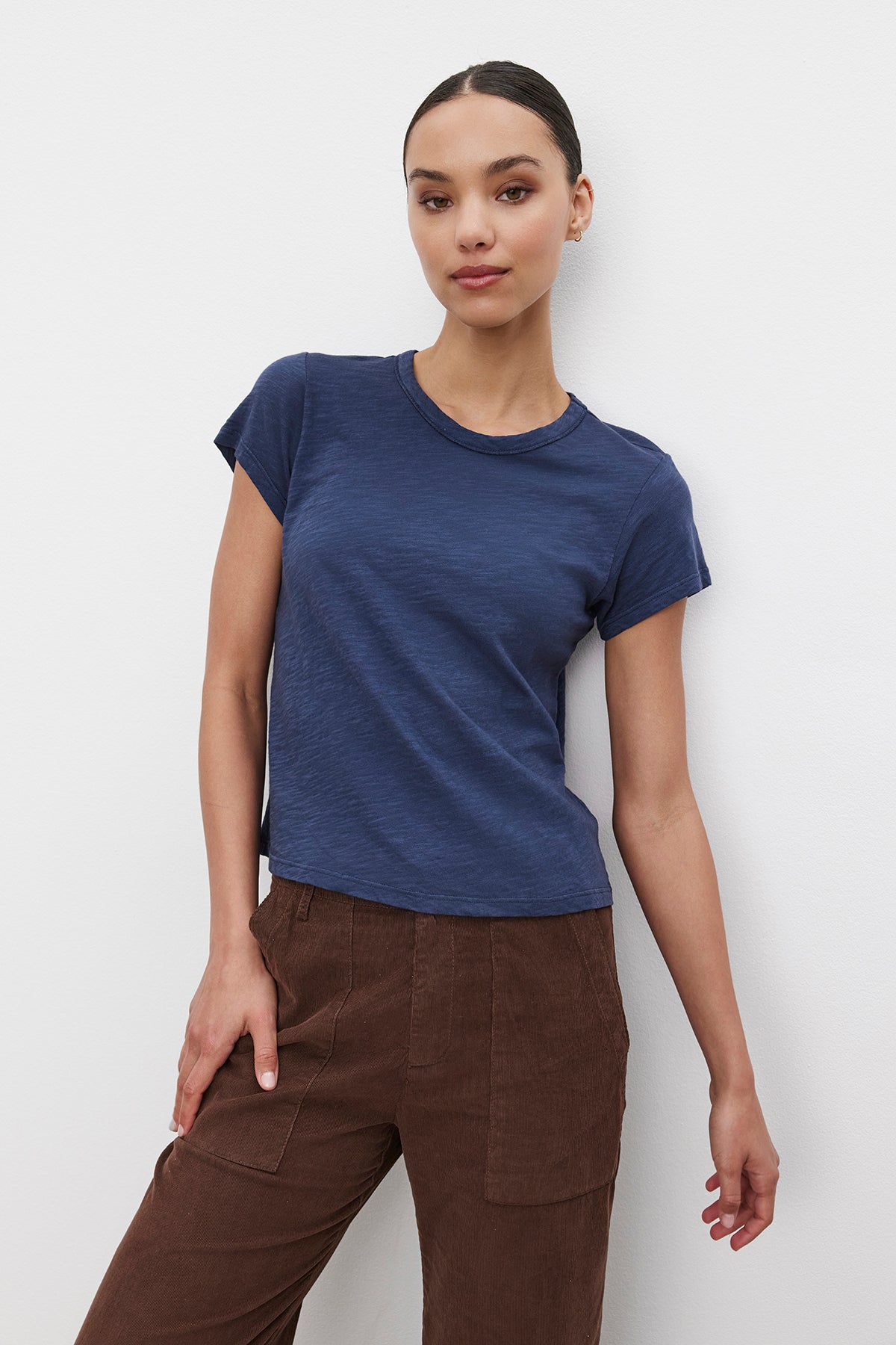   A person with dark hair tied back is wearing the MARIE TEE by Velvet by Graham & Spencer, a navy blue cotton slub T-shirt, paired with brown pants and standing against a plain white background. The outfit's boxy fit makes it perfect for a casual occasion. 