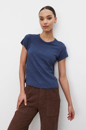 A person with dark hair tied back is wearing the MARIE TEE by Velvet by Graham & Spencer, a navy blue cotton slub T-shirt, paired with brown pants and standing against a plain white background. The outfit's boxy fit makes it perfect for a casual occasion.