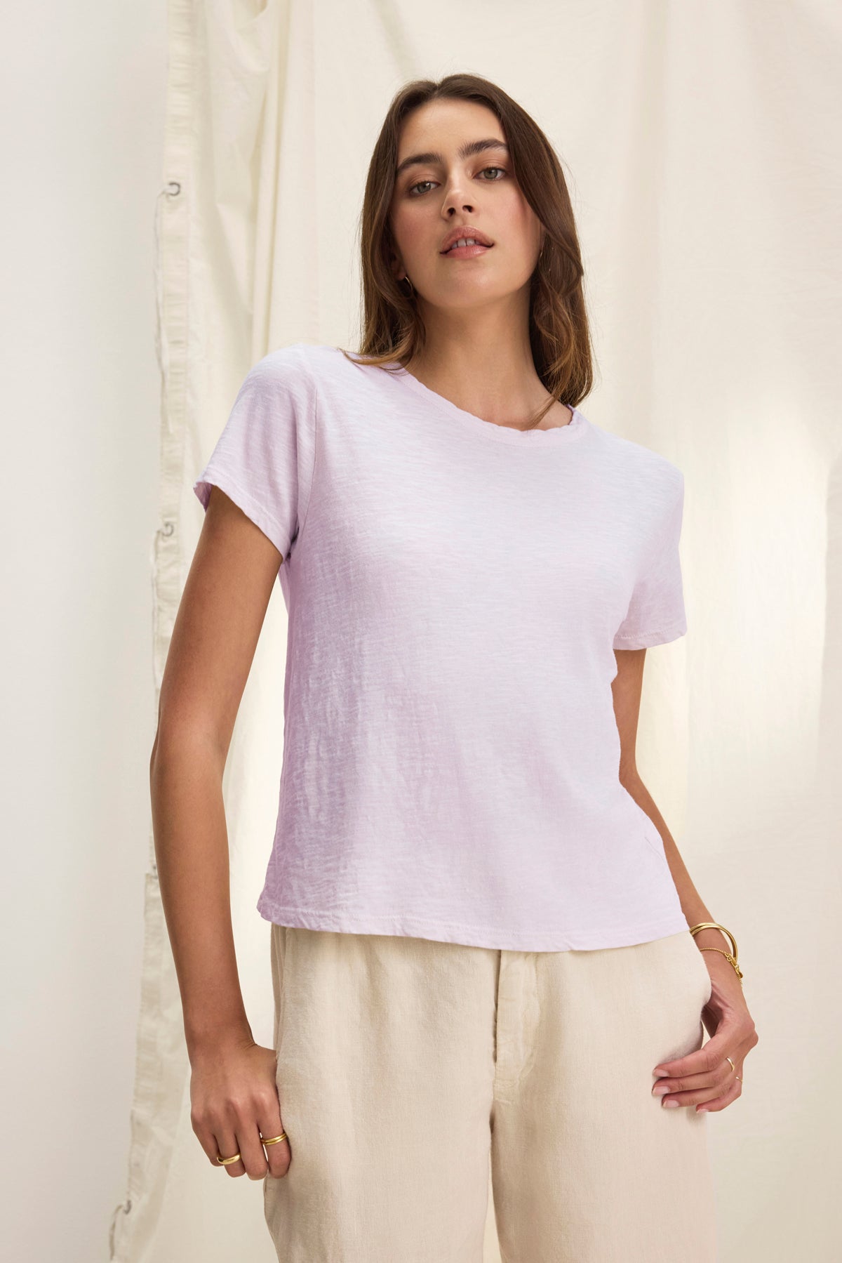   A person wearing the MARIE TEE from Velvet by Graham & Spencer, characterized by its light purple shade and relaxed boxy fit, paired with beige pants, stands in front of a cream-colored curtain. 