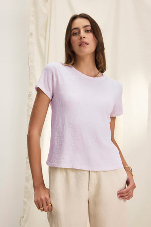 A person wearing the MARIE TEE from Velvet by Graham & Spencer, characterized by its light purple shade and relaxed boxy fit, paired with beige pants, stands in front of a cream-colored curtain.