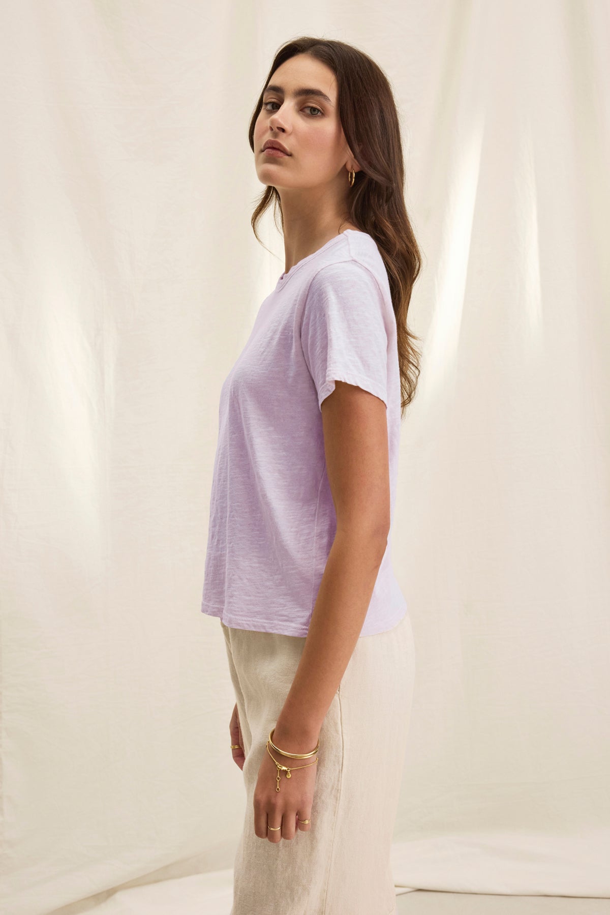   A woman stands sideways wearing the MARIE TEE by Velvet by Graham & Spencer in light purple, featuring a relaxed, boxy fit with beige pants against a neutral background. 