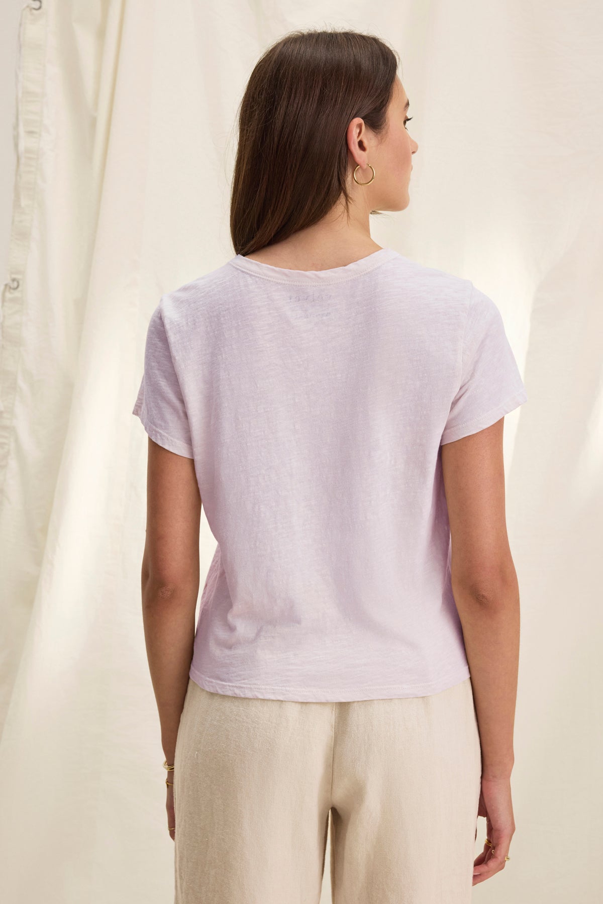   A long-haired brunette woman is seen from behind wearing the casual boxy-fit Velvet by Graham & Spencer MARIE TEE in light purple, paired with beige pants. A light fabric backdrop serves as the background. 