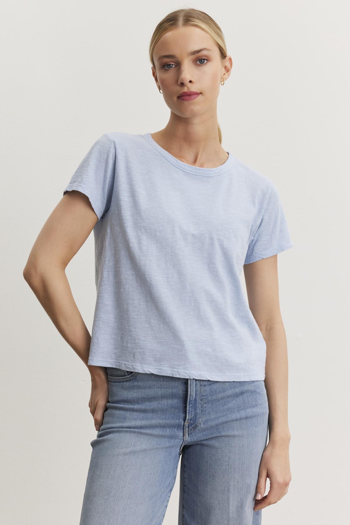   A person with blonde hair stands against a plain background, wearing a light blue MARIE TEE by Velvet by Graham & Spencer with a crew neckline and blue jeans. 