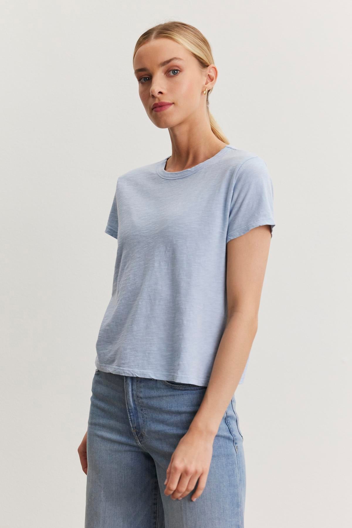   A person with their hair pulled back is wearing a light blue Velvet by Graham & Spencer MARIE TEE with a crew neckline and jeans, standing against a plain white background. 