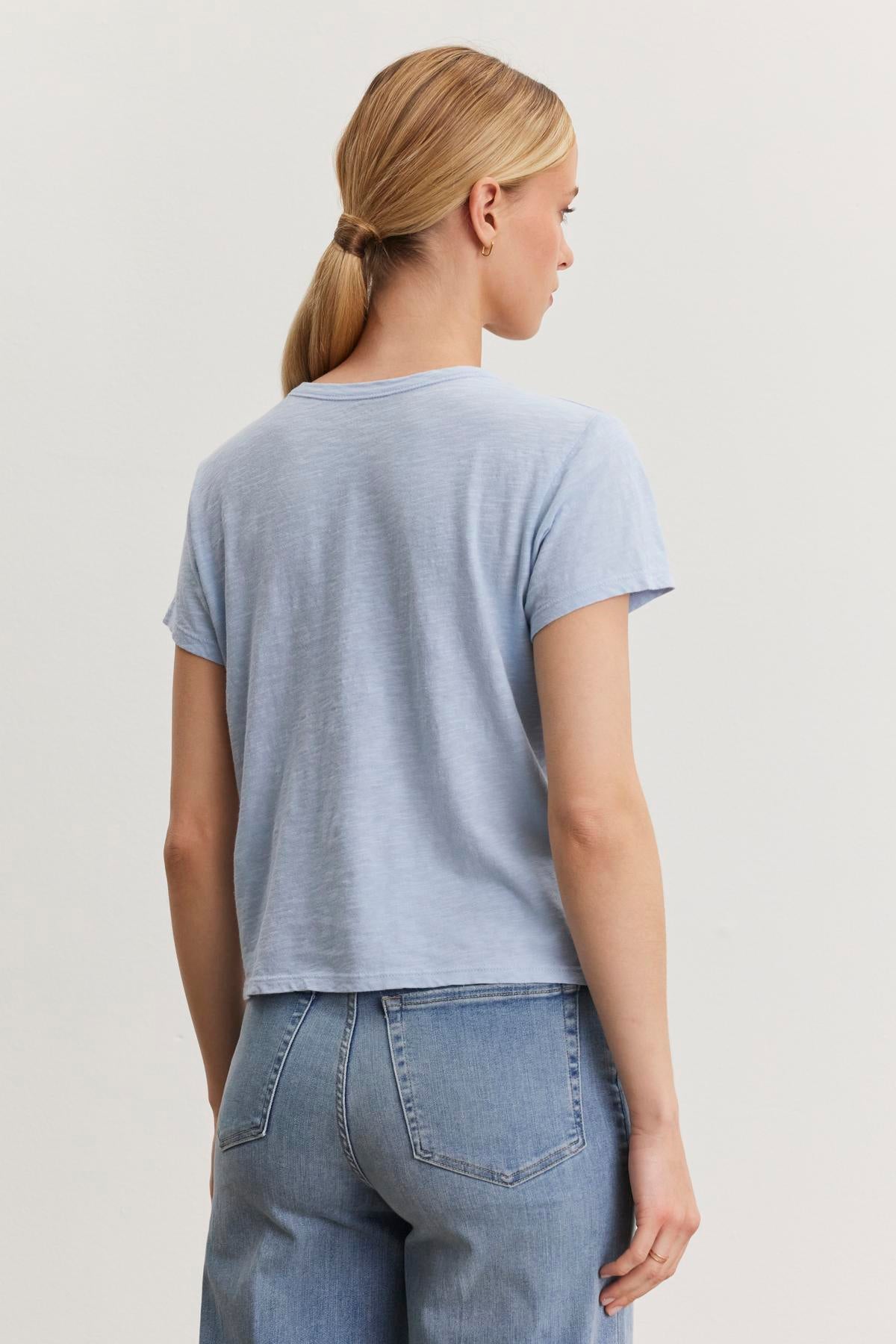   A person with blonde hair in a ponytail, wearing a light blue MARIE TEE by Velvet by Graham & Spencer with a crew neckline and relaxed boxy fit, paired with blue jeans, is facing away from the camera. 