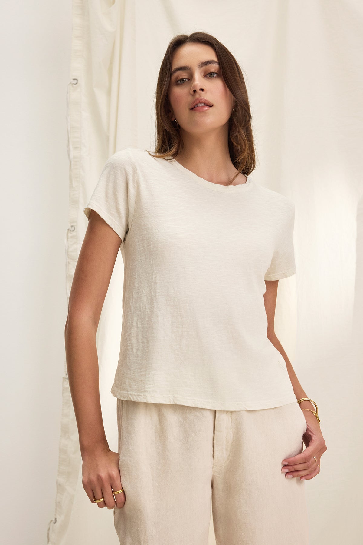   A woman in a white MARIE TEE by Velvet by Graham & Spencer, featuring a relaxed boxy fit and crew neckline, pairs it effortlessly with beige pants against a light curtain backdrop. 