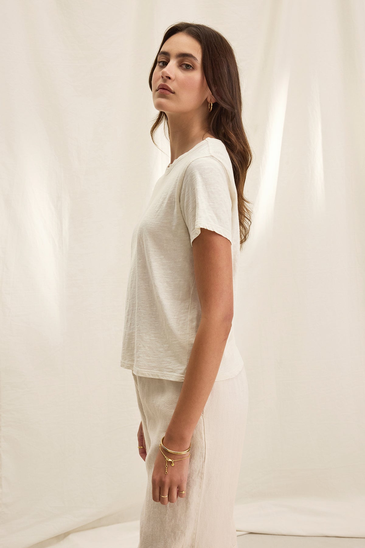   A woman is seen standing sideways in a relaxed, boxy fitted MARIE TEE by Velvet by Graham & Spencer, featuring a crew neckline paired with light-colored pants against a cream background. 