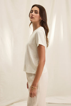 A woman is seen standing sideways in a relaxed, boxy fitted MARIE TEE by Velvet by Graham & Spencer, featuring a crew neckline paired with light-colored pants against a cream background.