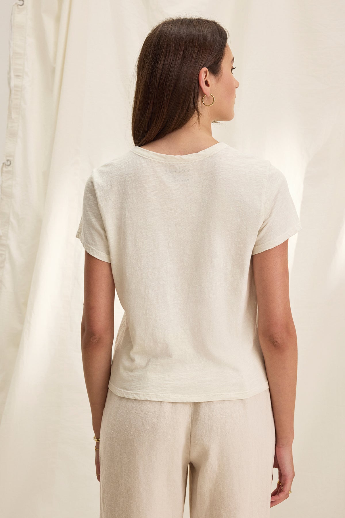   A woman with long hair stands with her back to the camera, wearing the MARIE TEE from Velvet by Graham & Spencer. The light beige T-shirt has a crew neckline and features a relaxed boxy fit, paired with pants, all set against a cream-colored backdrop. 