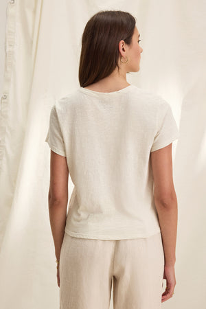 A woman with long hair stands with her back to the camera, wearing the MARIE TEE from Velvet by Graham & Spencer. The light beige T-shirt has a crew neckline and features a relaxed boxy fit, paired with pants, all set against a cream-colored backdrop.