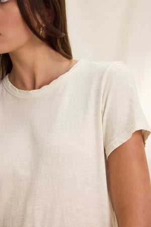 A person wearing a MARIE TEE from Velvet by Graham & Spencer, with a beige boxy fit and gold hoop earrings, stands against a neutral background, perfect for casual occasions.