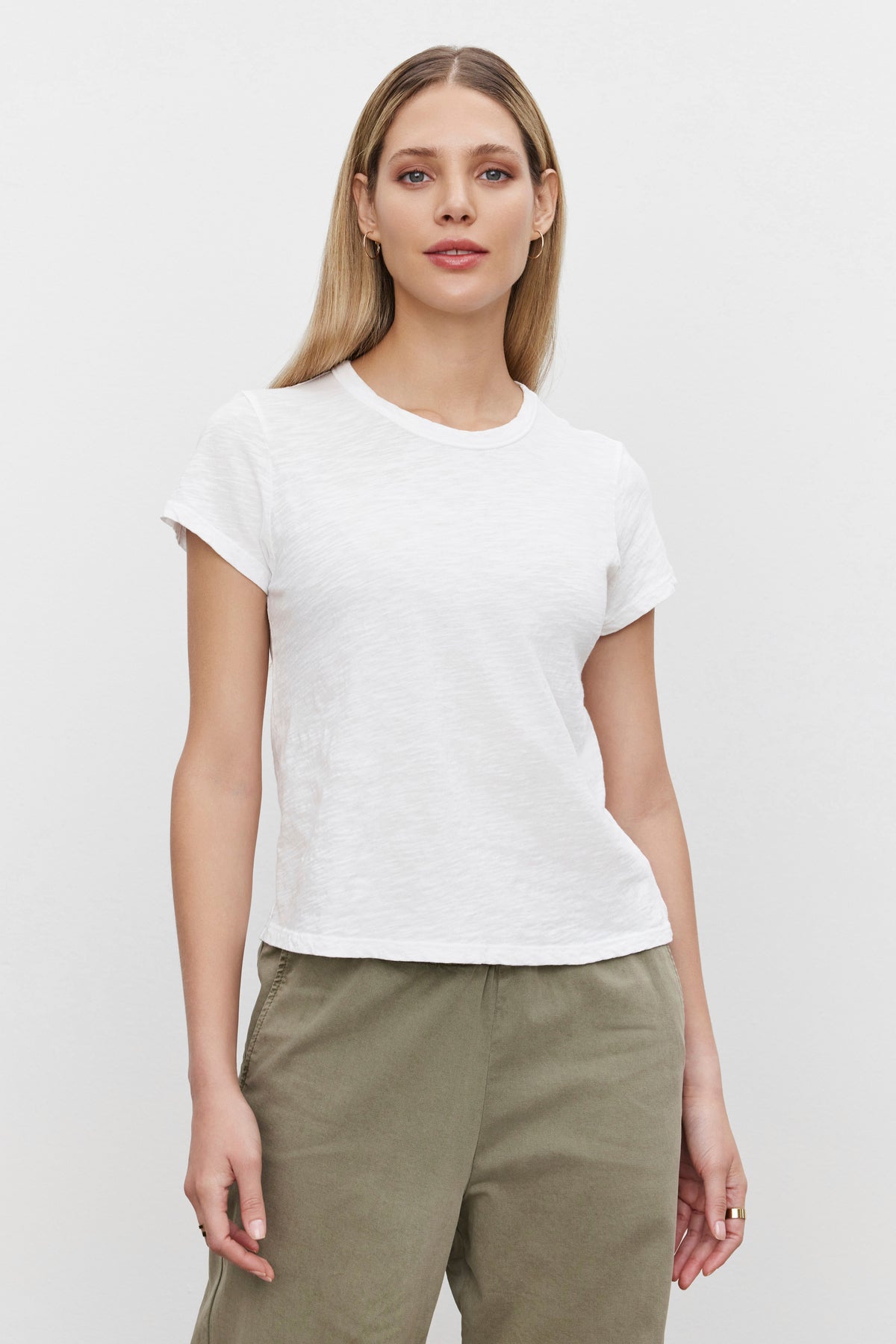   A person with long hair is wearing the MARIE TEE, a plain white cotton slub T-shirt from Velvet by Graham & Spencer, paired with olive green pants. They stand against a plain white background. Perfect for a casual occasion, the relaxed boxy fit of the MARIE TEE adds to their laid-back look. 