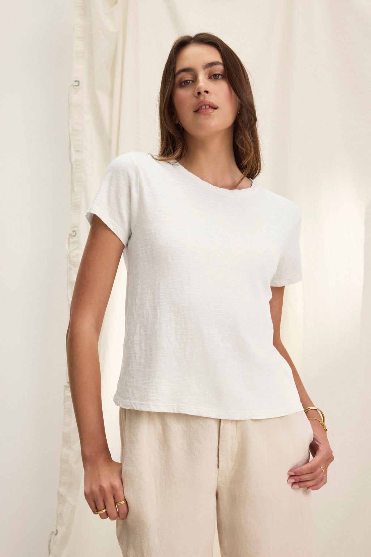   A person stands against a light curtain, wearing the Velvet by Graham & Spencer MARIE TEE in plain white and beige pants. Their long brown hair cascades down as they look directly at the camera. 