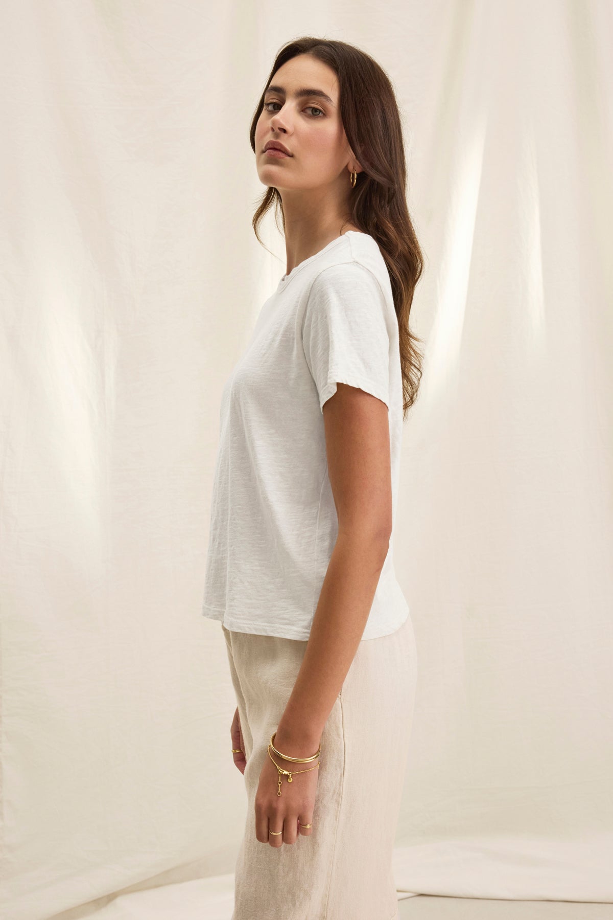   A woman in a casual MARIE TEE and beige pants stands sideways against a fabric backdrop. Her long brown hair cascades down as she accessorizes with hoop earrings and bracelets, embracing a relaxed style perfect for everyday ease. Brand: Velvet by Graham & Spencer. 