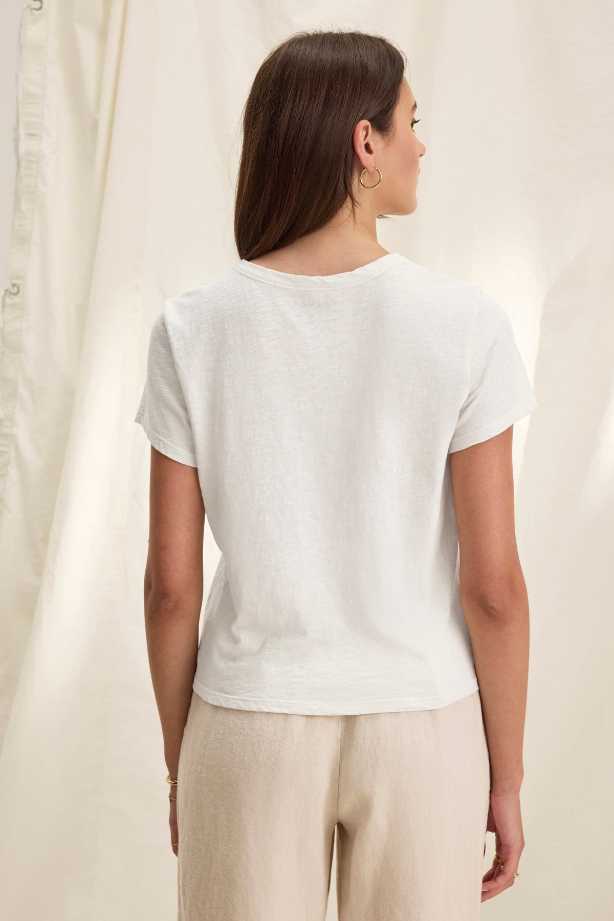   A person with long brown hair faces away, wearing the relaxed fit MARIE TEE by Velvet by Graham & Spencer and beige pants, set against a light, draped fabric backdrop. 