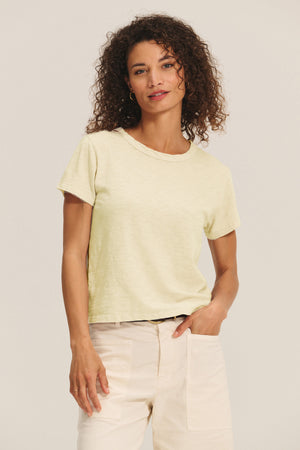 A woman with curly hair wears the MARIKA TEE in light yellow by Velvet by Graham & Spencer—a cotton slub boxy-fit top—paired with beige pants, standing against a plain background.