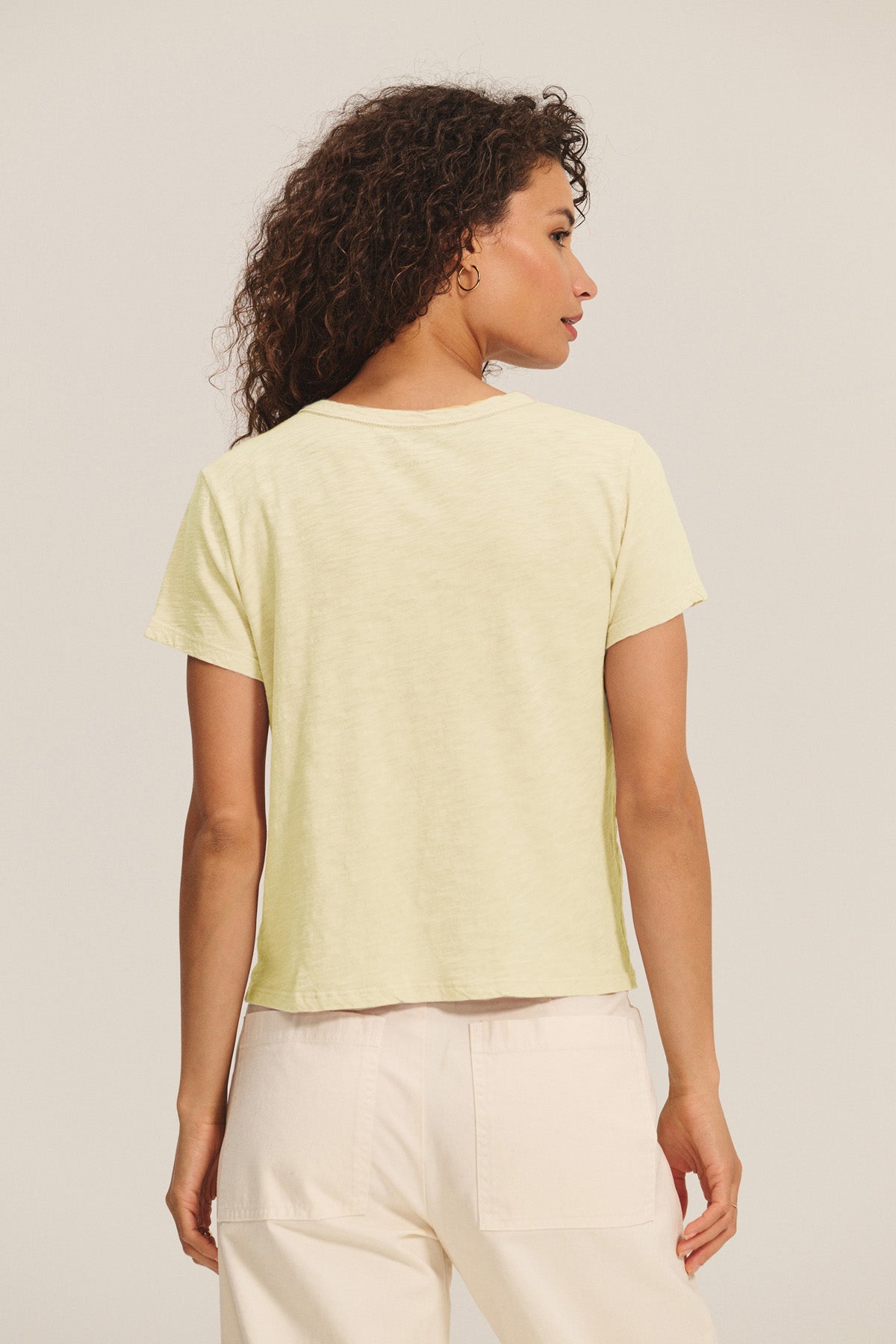 A woman with curly hair is seen from the back wearing a light yellow MARIKA TEE by Velvet by Graham & Spencer in a relaxed boxy fit, paired with light pants, set against a plain background.-40680354480321