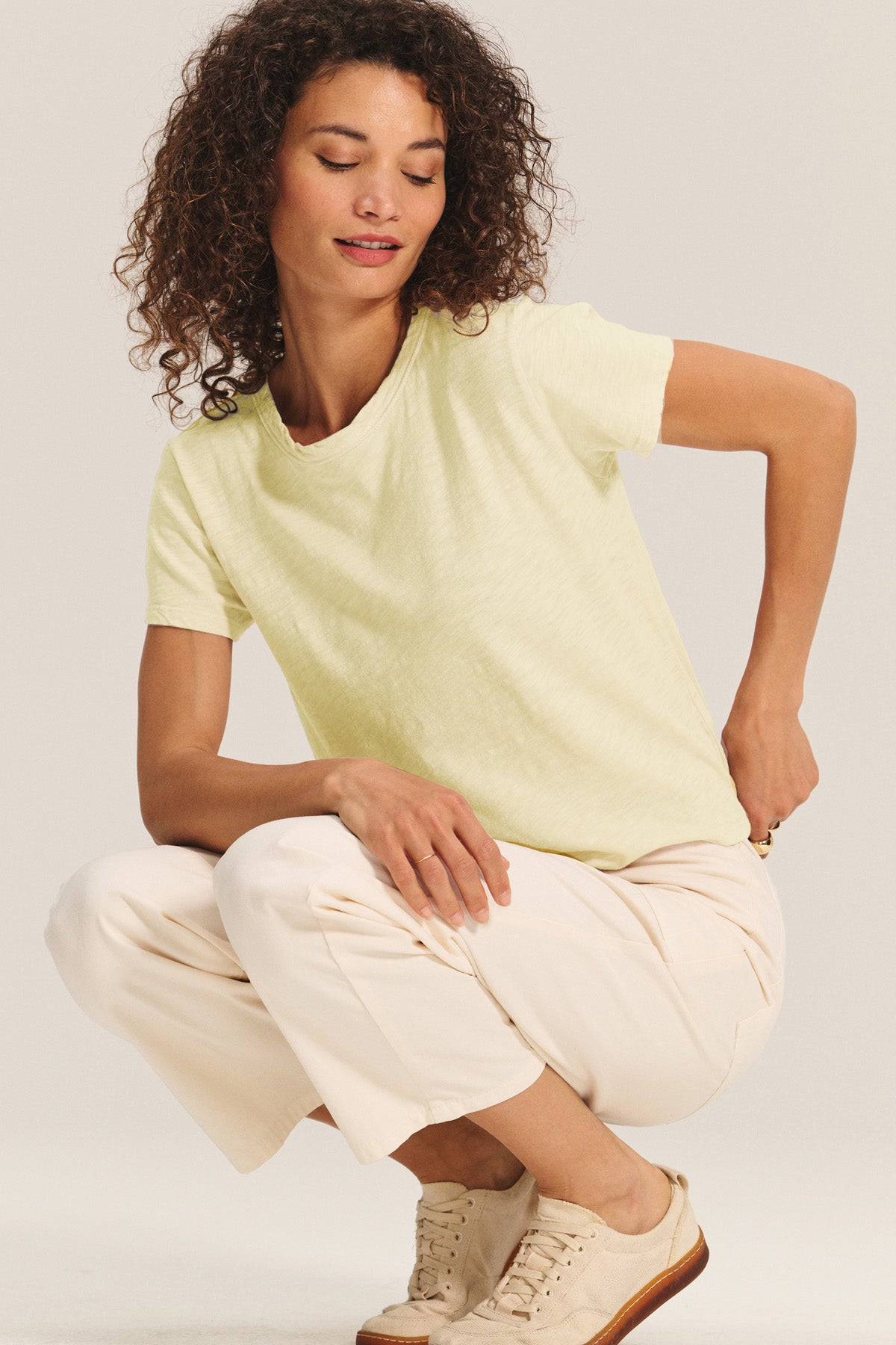 Woman with curly hair wears a relaxed boxy fit MARIKA TEE in pale yellow cotton slub by Velvet by Graham & Spencer with white pants and beige sneakers, crouching on one knee against a plain light background.-40680354447553