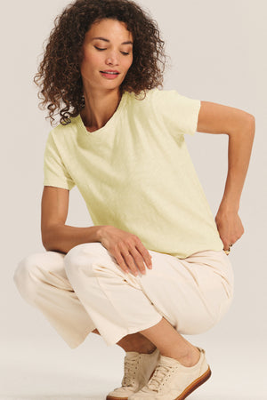 Woman with curly hair wears a relaxed boxy fit MARIKA TEE in pale yellow cotton slub by Velvet by Graham & Spencer with white pants and beige sneakers, crouching on one knee against a plain light background.