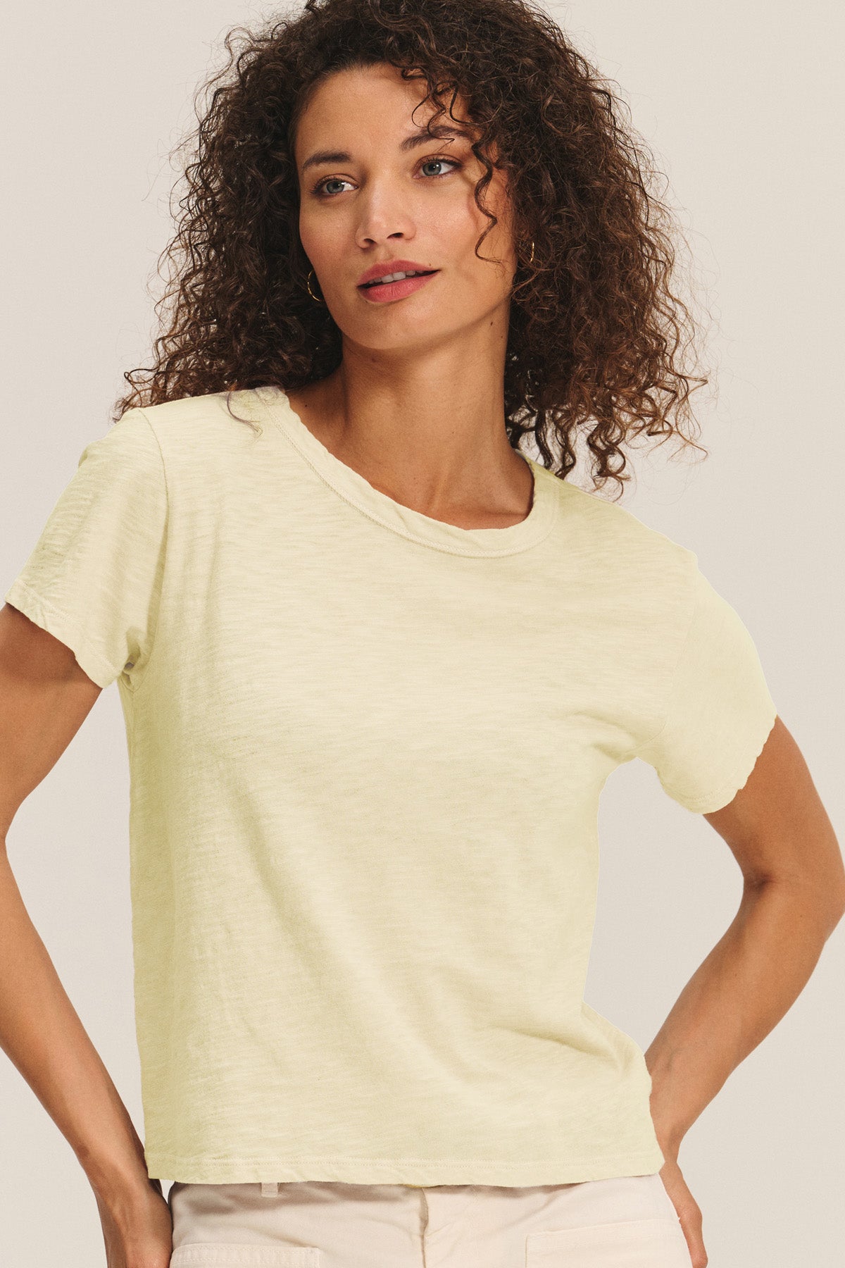 A woman with curly hair wears the MARIKA TEE in light yellow cotton slub by Velvet by Graham & Spencer and beige pants, showing its relaxed boxy fit against a neutral background.-40680354414785