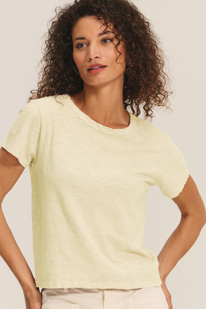 A woman with curly hair wears the MARIKA TEE in light yellow cotton slub by Velvet by Graham & Spencer and beige pants, showing its relaxed boxy fit against a neutral background.