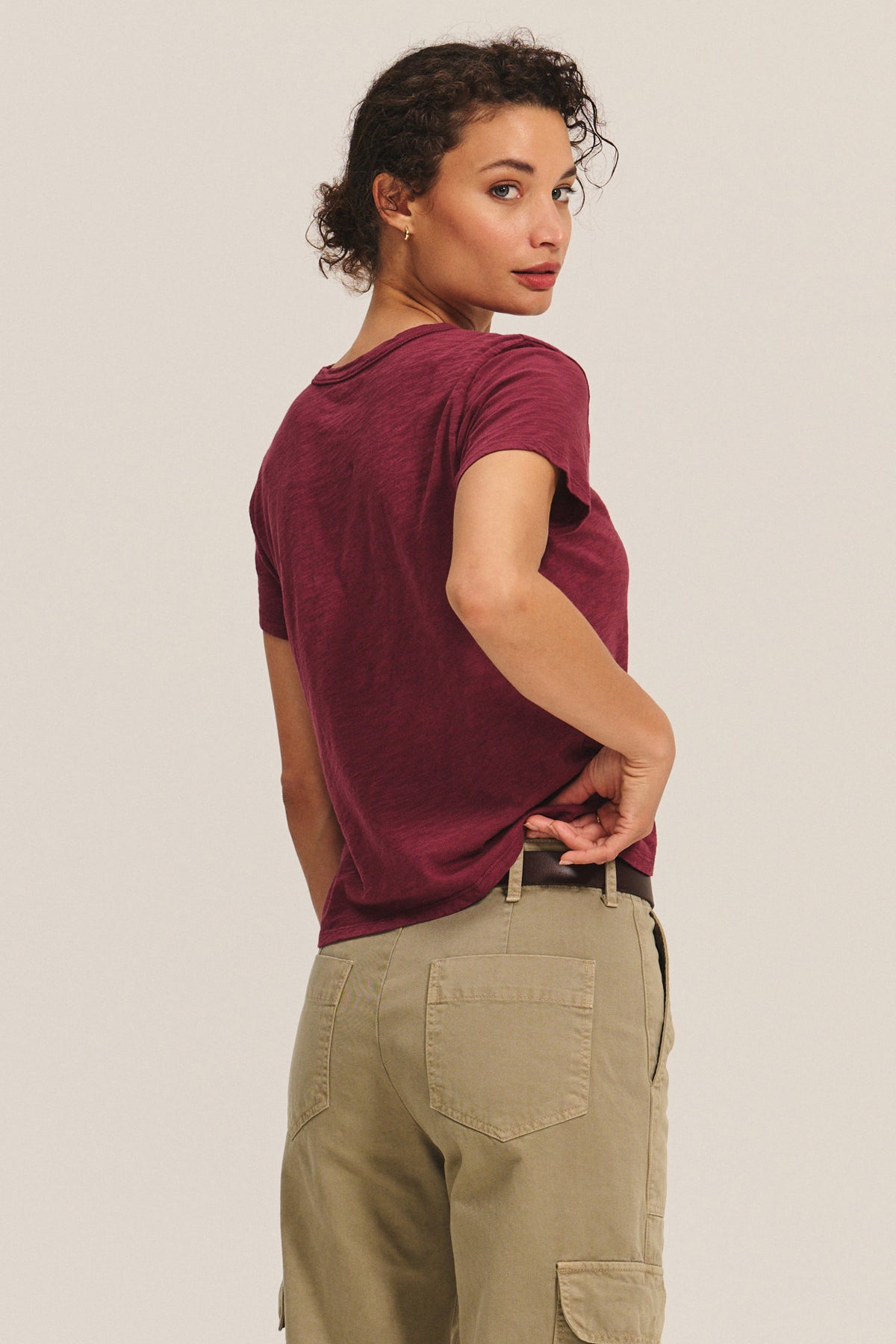   A person with short curly hair, in a relaxed boxy-fit maroon MARIKA TEE by Velvet by Graham & Spencer and beige cargo pants, stands facing away and looks over their shoulder. 