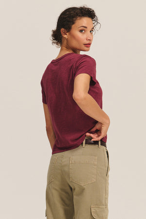 A person with short curly hair, in a relaxed boxy-fit maroon MARIKA TEE by Velvet by Graham & Spencer and beige cargo pants, stands facing away and looks over their shoulder.