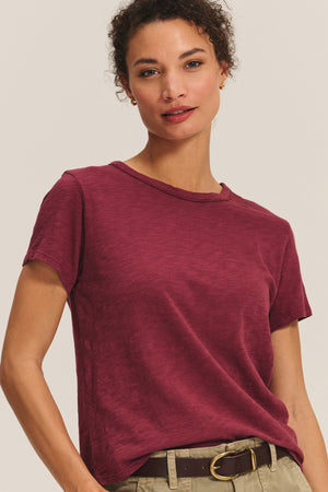 A person wears a short-sleeved, maroon MARIKA TEE by Velvet by Graham & Spencer in a relaxed boxy fit, paired with khaki pants and a brown belt, standing against a plain background.