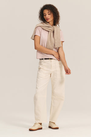 Wearing a boxy fit MARIKA TEE in pink by Velvet by Graham & Spencer, cream wide-leg pants, and a beige sweater over the shoulders, a person stands against a plain background.