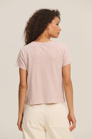 A woman with curly hair is shown from the back, wearing a light pink MARIKA TEE by Velvet by Graham & Spencer in soft cotton slub and off-white pants against a plain background.