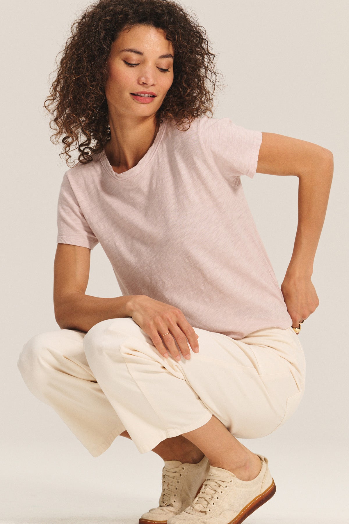 A woman with curly hair wears a light pink MARIKA TEE by Velvet by Graham & Spencer, paired with white pants and beige sneakers, squatting against a neutral background.-40598530031809