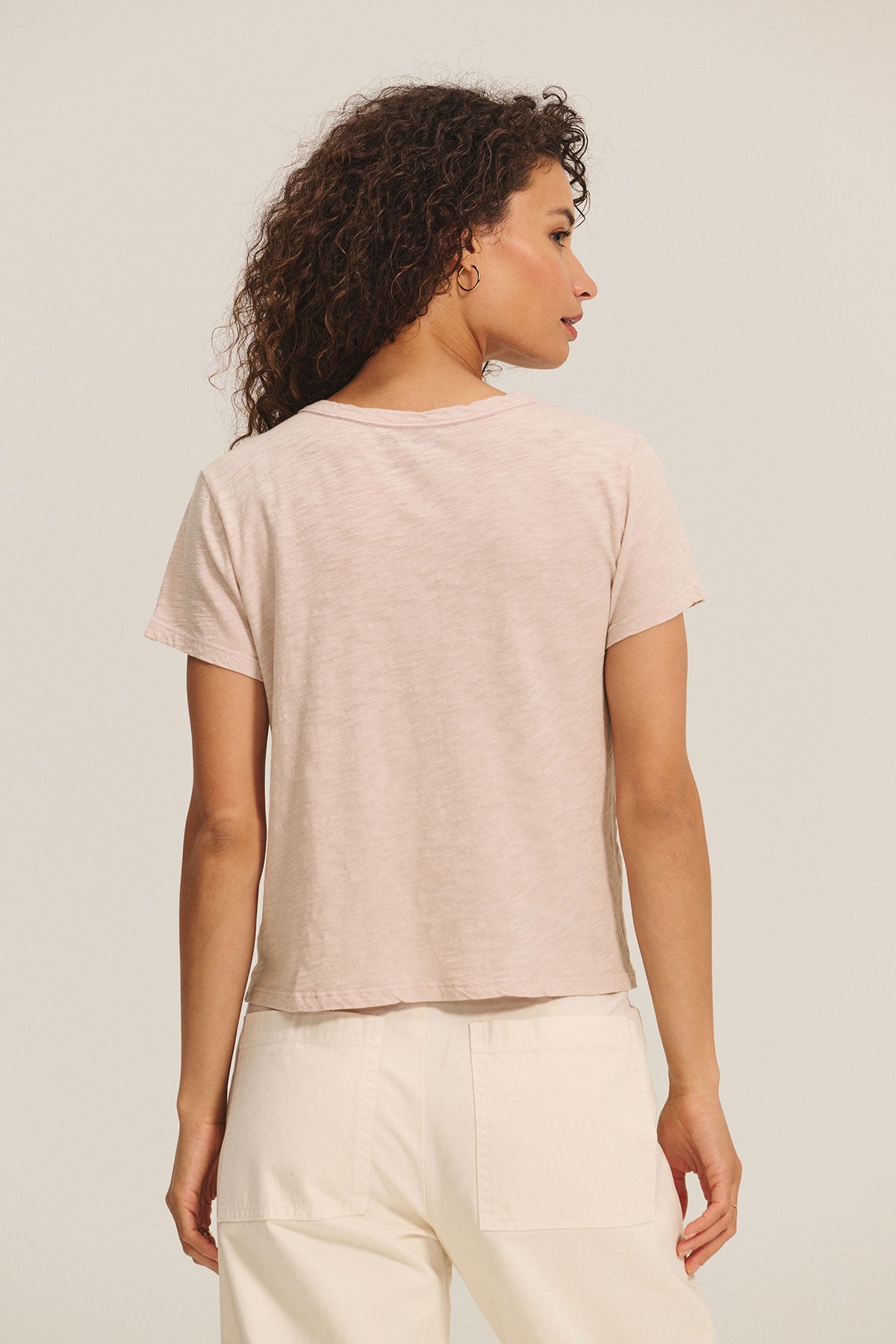 A woman with curly brown hair, shown from the back against a plain background, wears the MARIKA TEE by Velvet by Graham & Spencer in light pink cotton slub boxy fit with cream pants.